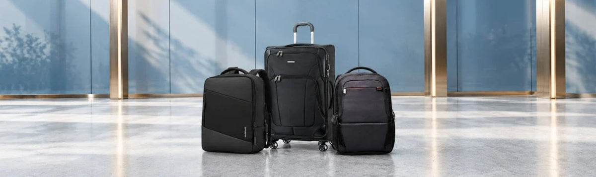 Black Samsonite softside suitcase and two business backpacks displayed in a modern atrium with blue glass walls and gold accents.