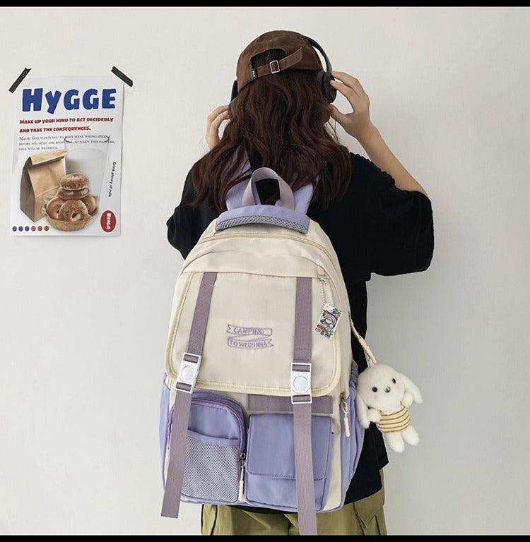 Beige and purple student backpack with front pockets and plush charm, worn by person with headphones