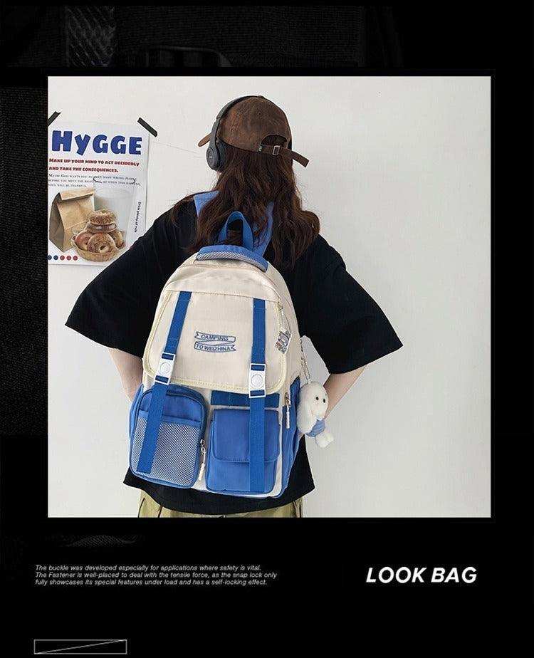 Blue and cream student backpack with front pockets and plush charm, worn by person with cap and headphones