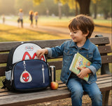 Child sitting on a park bench with a backpack and book, surrounded by trees and people in the background.